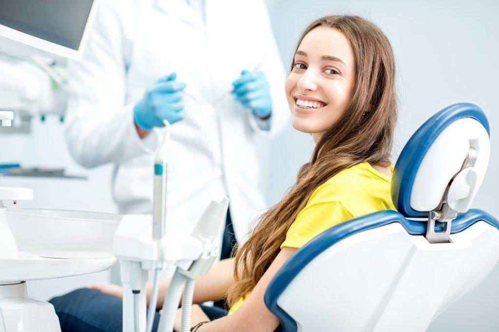 portrait-woman-with-toothy-smile-sitting-dental-chair-with-doctor-background-dental-office-scaled