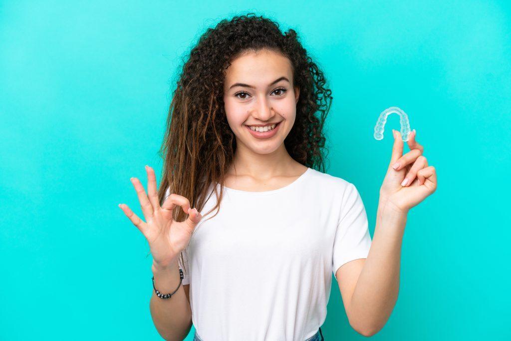 young-arab-woman-holding-invisible-braces-isolated-blue-background-showing-ok-sign-with-fingers-scaled
